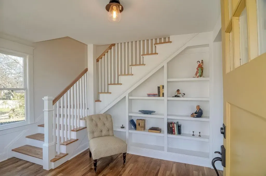 Bright hallway with white staircase, built-in shelves, beige chair, and warm wood floors