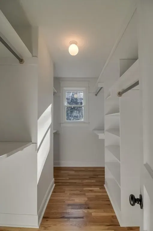 Narrow white walk-in closet with built-in shelves, hanging rods, wood floor, and a small window.