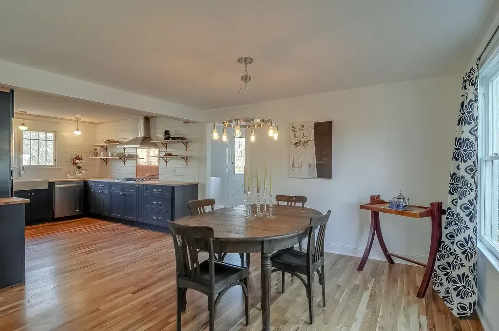 Bright dining area with wooden table, dark chairs, open kitchen, and large window curtains