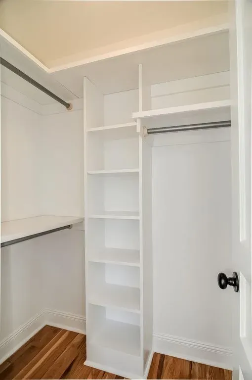 Empty white walk-in closet with built-in shelving, hanging rods, and hardwood floor