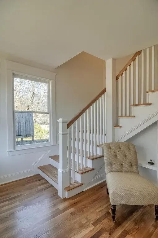 Bright foyer with wood floors, white staircase, large window, and a beige upholstered chair.