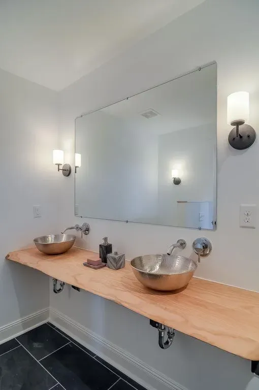 Bathroom vanity with large mirror, wall sconces, two metal sinks, and a wooden countertop with toiletries