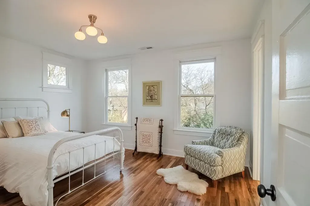 Bright white bedroom with metal bed, two windows, patterned chair, and hardwood floor