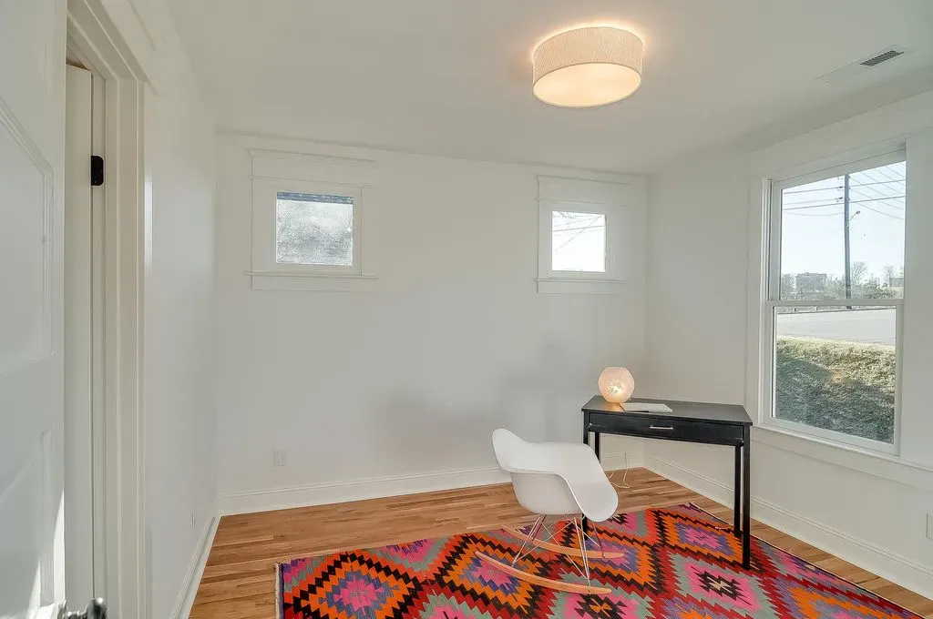 Bright white room with a desk, chair, colorful rug, and windows letting in daylight.