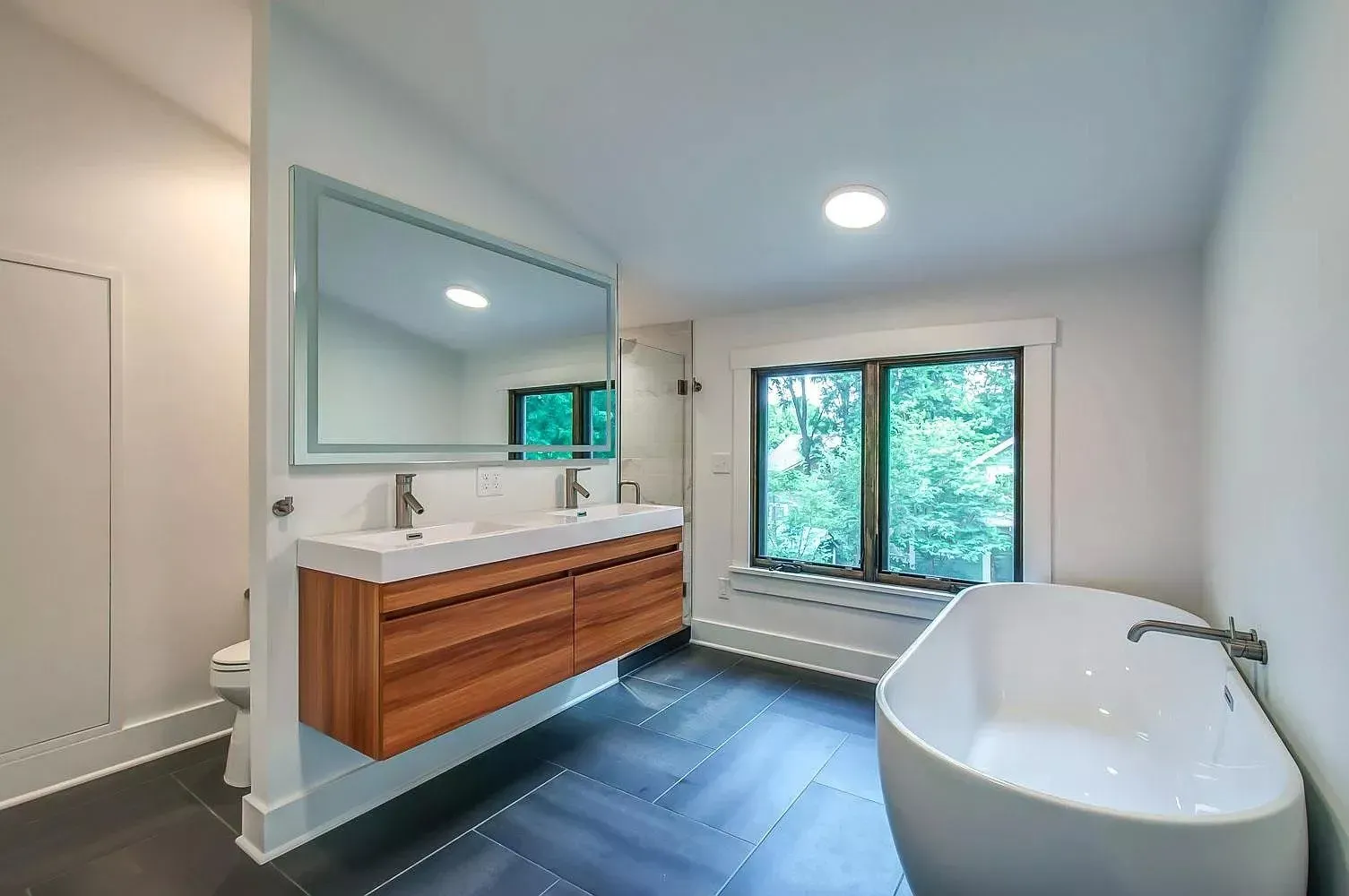 Modern bathroom with floating wood vanity, large mirror, and freestanding tub by a window