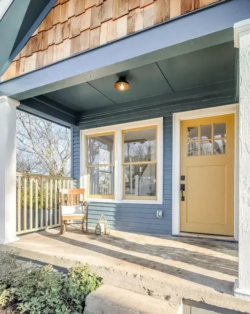 Front porch with yellow door, windows, blue siding, and a small bench in sunlight