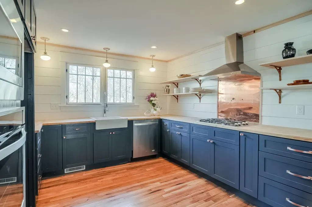 Modern kitchen with navy cabinets, wood floors, white walls, stainless range hood, and a window over the sink