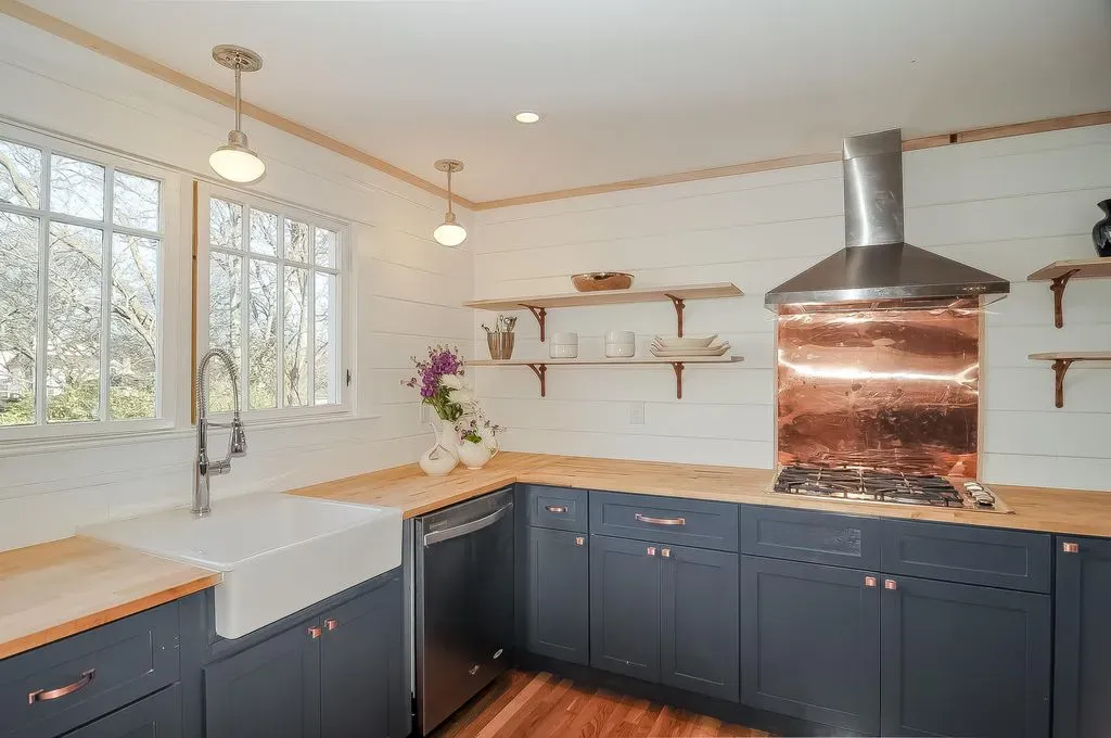 Bright kitchen with navy cabinets, white sink, wood countertops, and a copper range hood by large windows