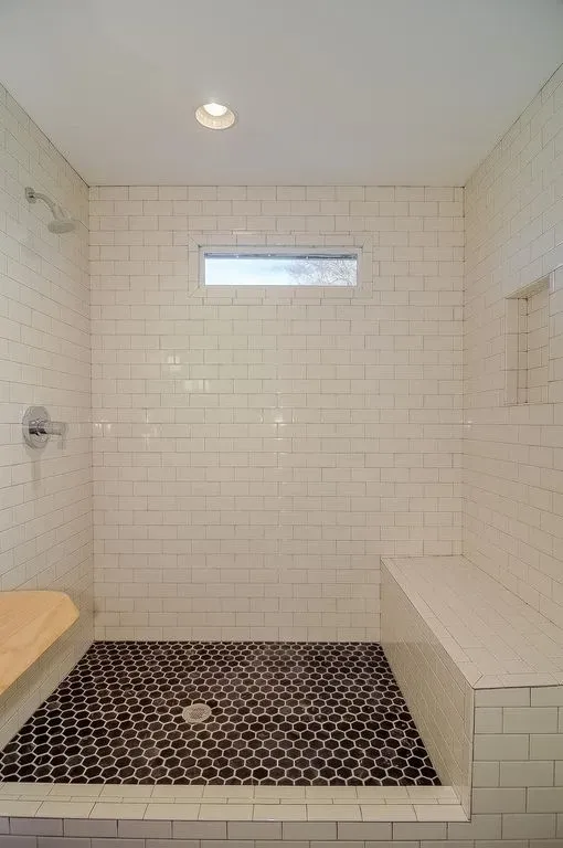 Empty white-tiled walk-in shower with a small window, dark hexagonal floor tiles, and a bench.