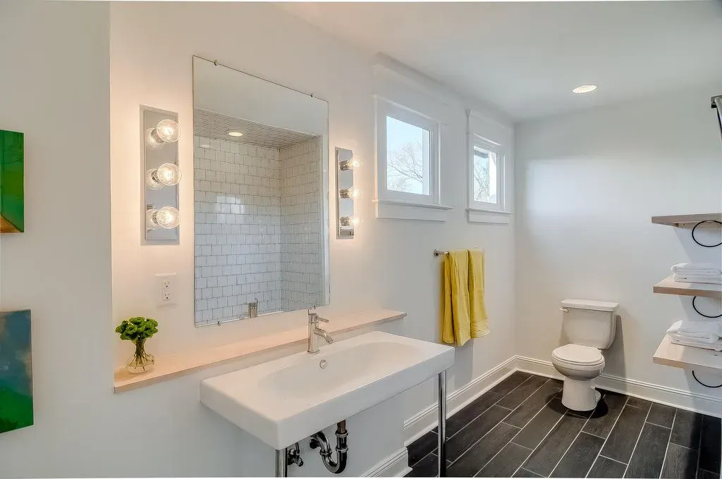 Bright white bathroom with double vanity, large mirror, yellow towels, toilet, and dark floor tiles.