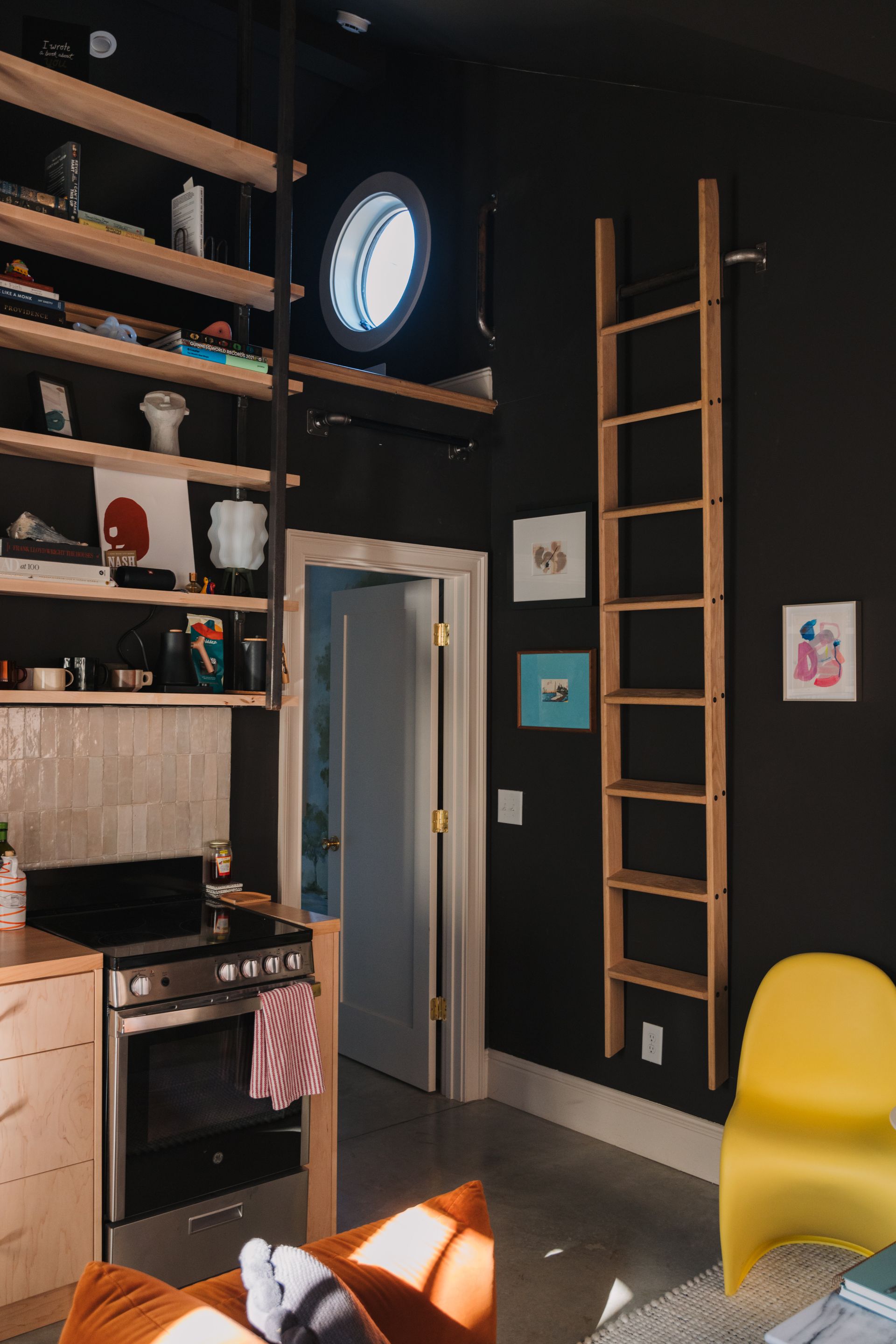 Modern kitchen with black walls, wooden shelves, ladder, yellow chair, and sunlight on the floor