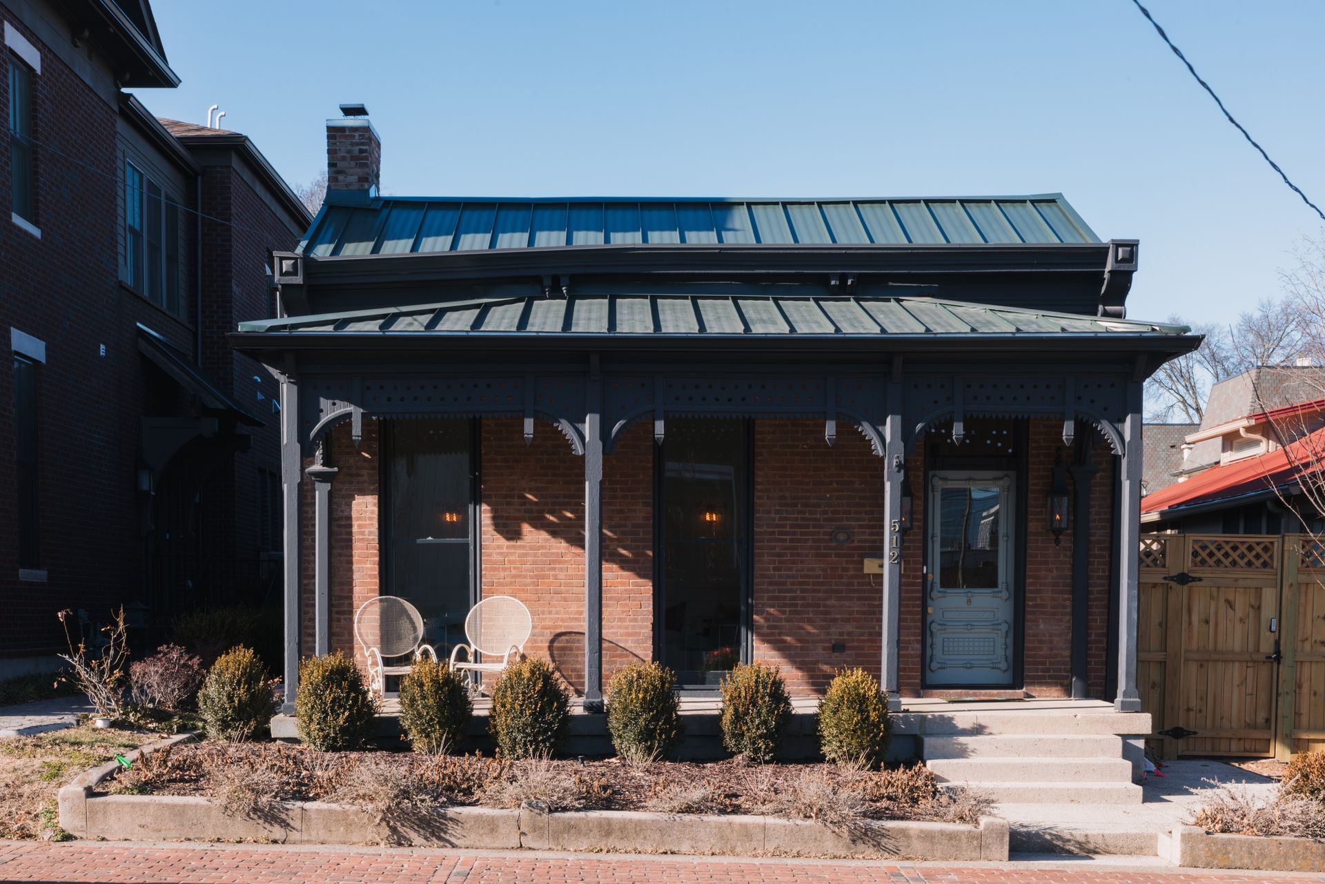 Brick house with a front porch, black trim, potted shrubs, and a wooden fence on a sunny day