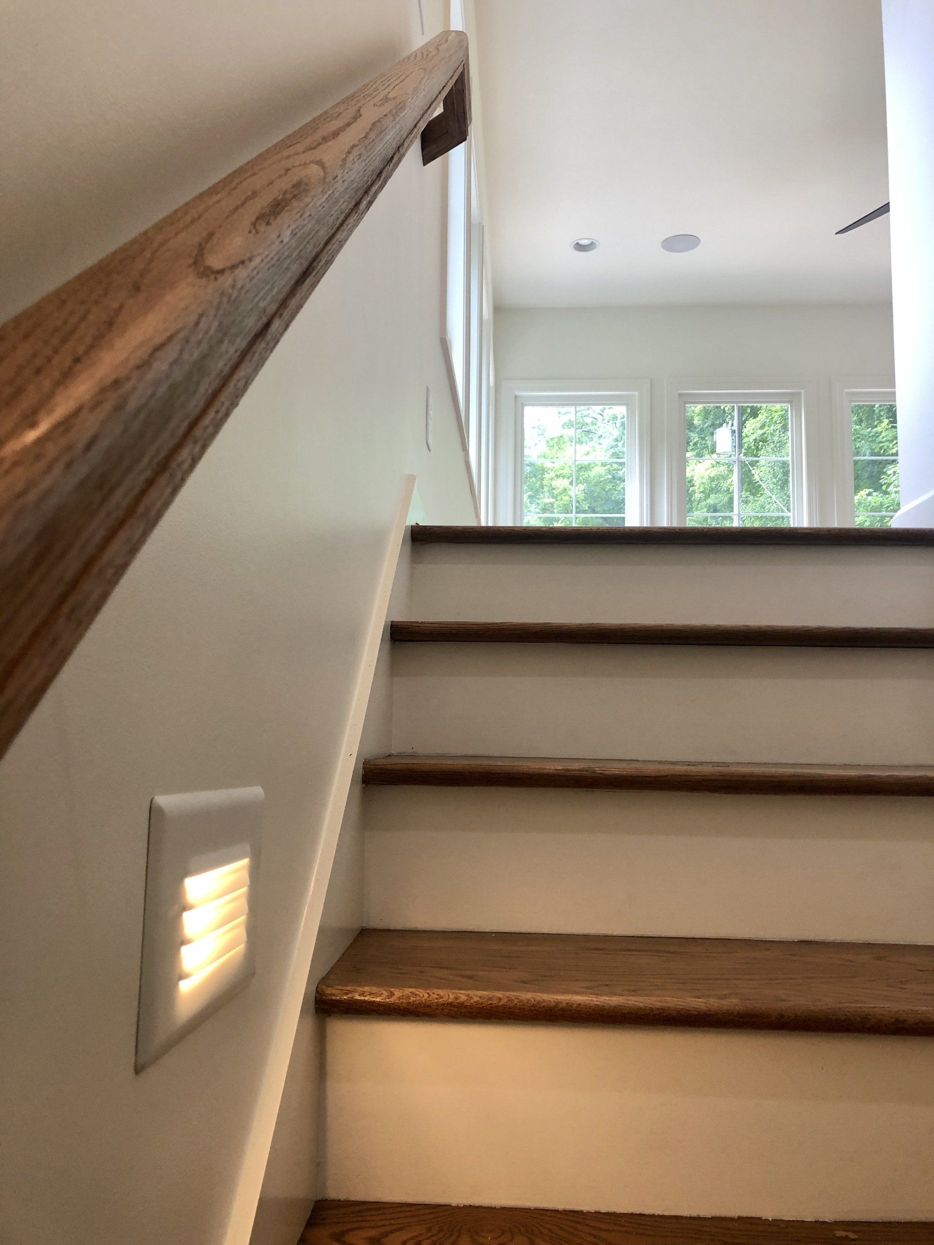 Interior staircase with wooden handrail and steps, lit by a wall light, leading to a bright windowed landing