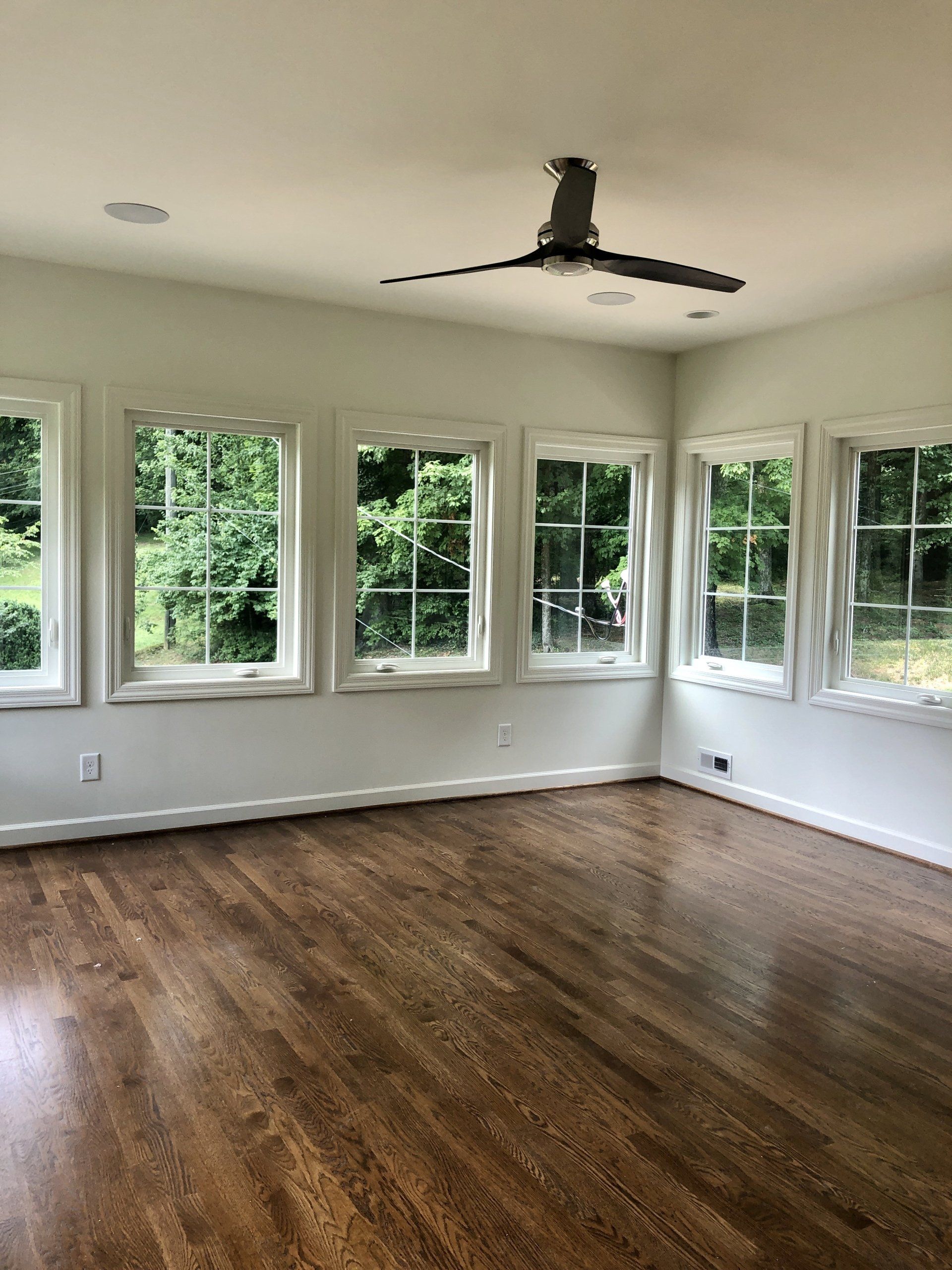 Empty room with hardwood floors, white walls, and large corner windows overlooking greenery