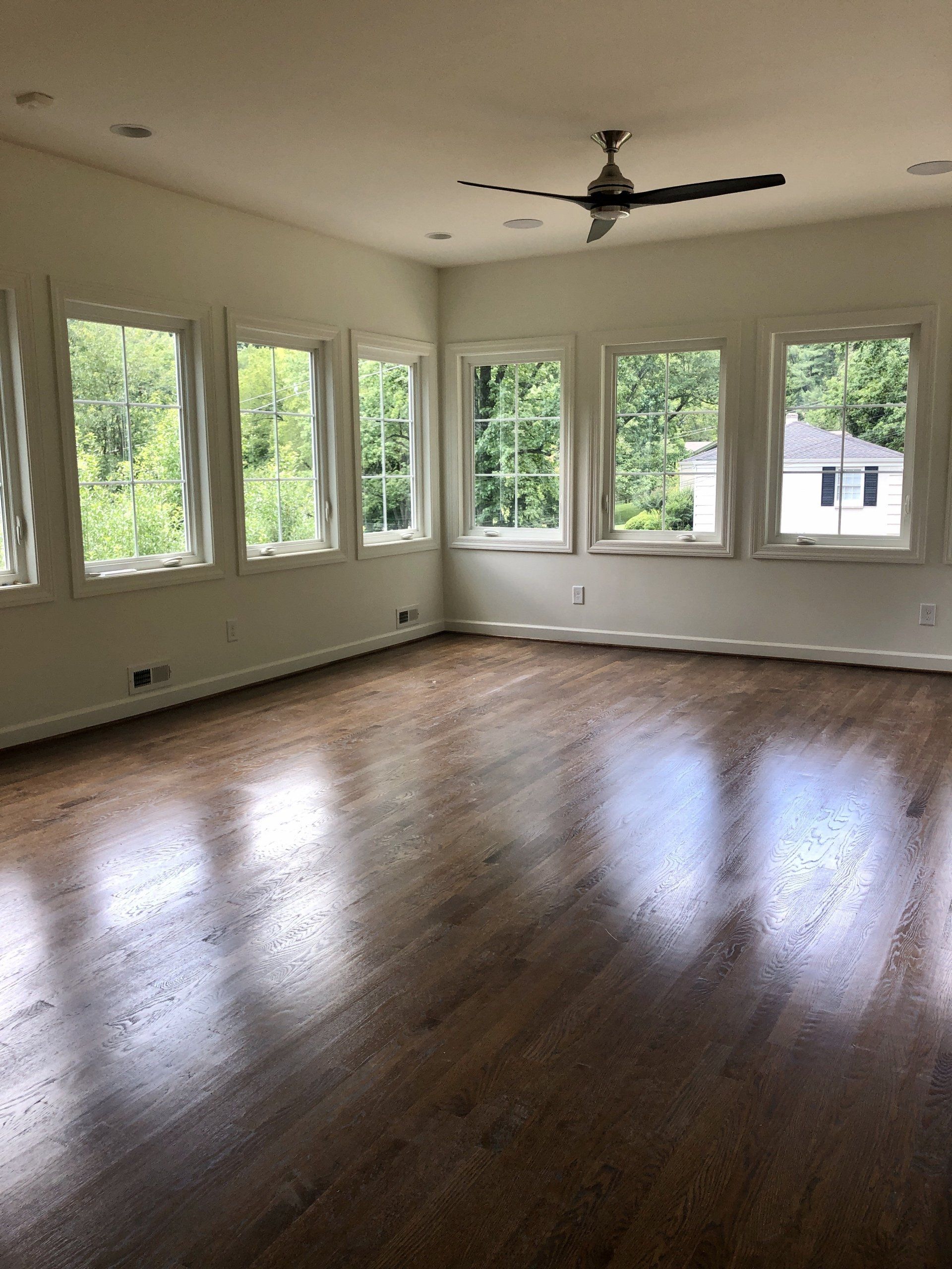 Empty bright room with hardwood floors, large windows, and a ceiling fan.