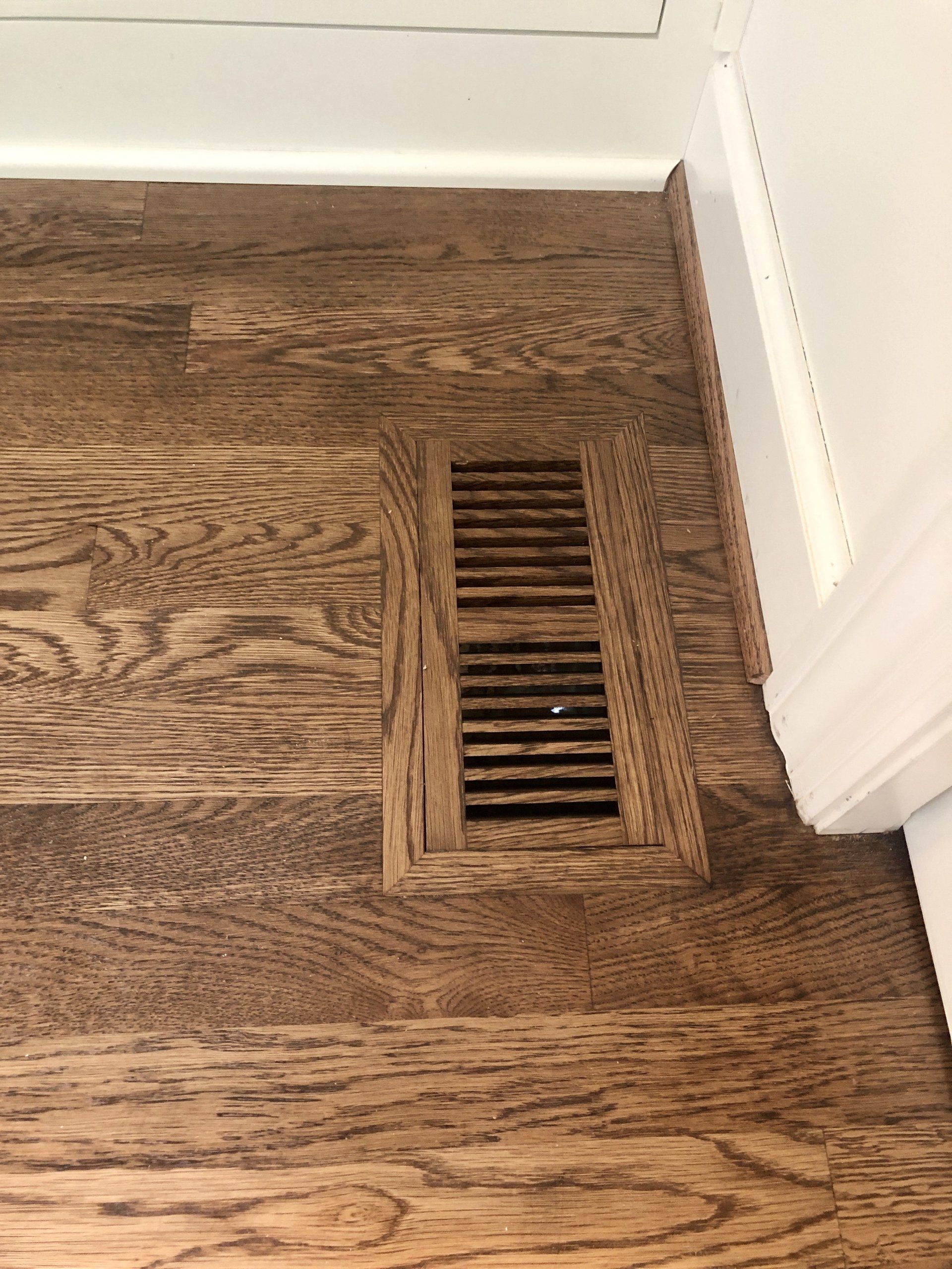 Wood floor air vent beside a white baseboard and cabinet in a corner.