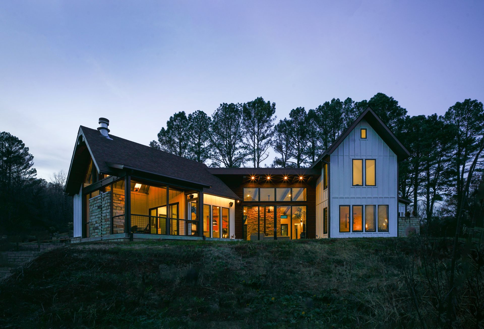 Modern house with warm lights at dusk, set on a grassy hill against a line of tall trees