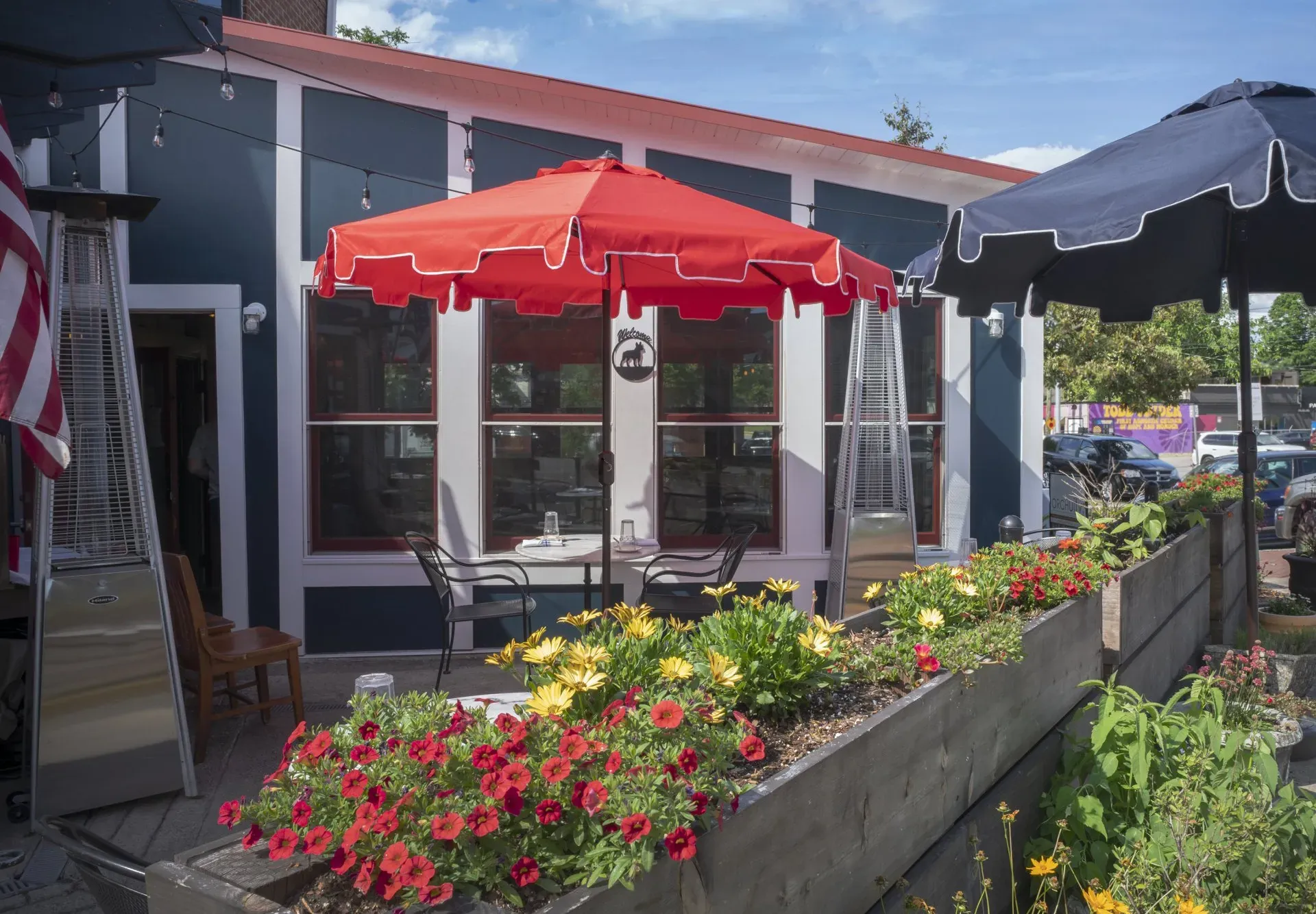 Outdoor cafe with red and black umbrellas, flower boxes, and storefront seating on a sunny day