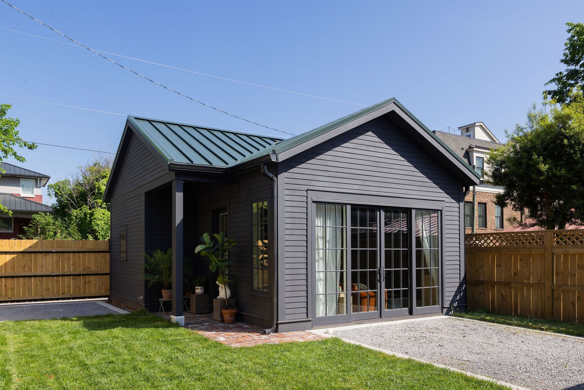 Small gray house with a green metal roof, large windows, and a fenced yard under a clear blue sky