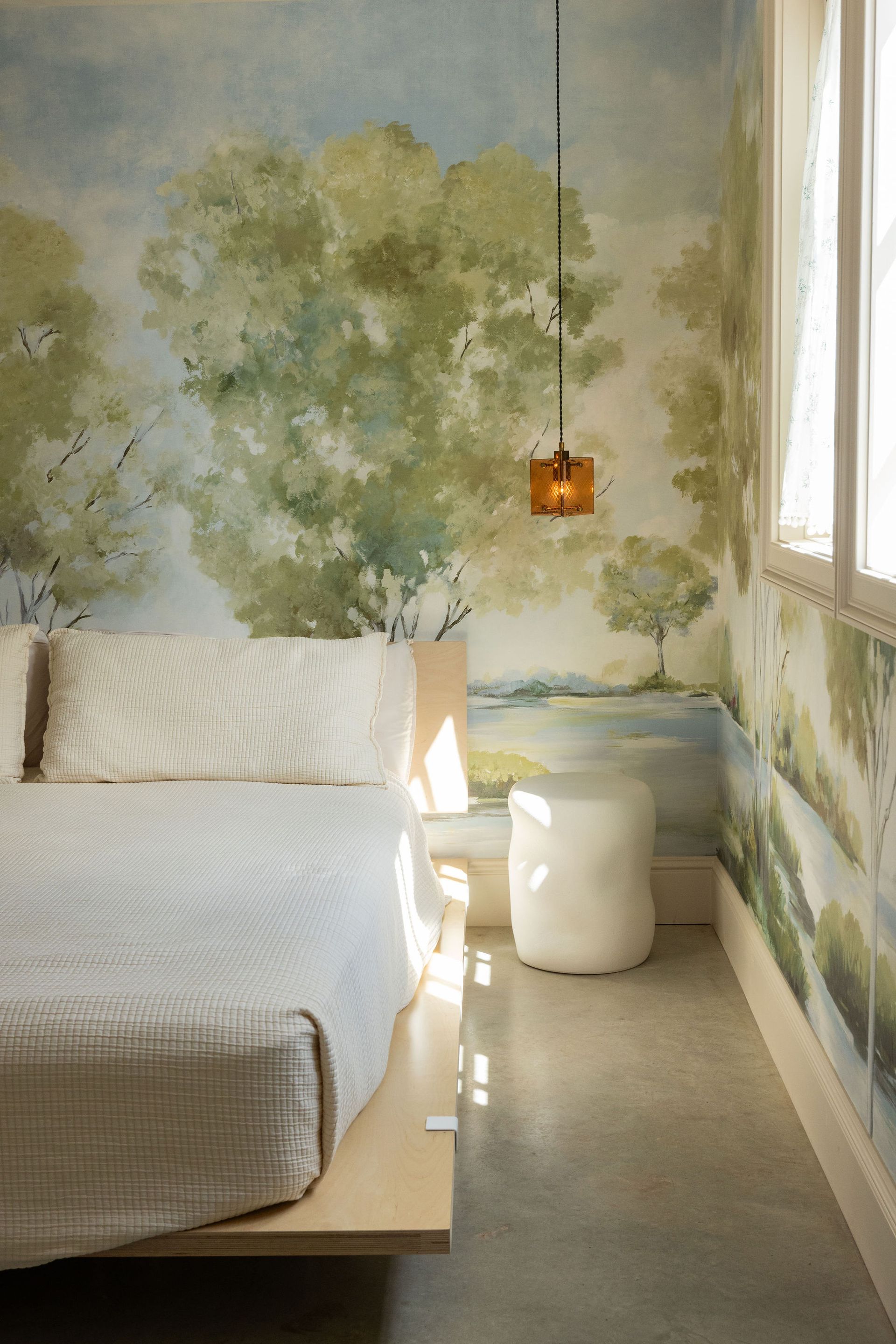 Cozy bedroom with bed, leafy mural wall, hanging lamp, and sunlight by the window