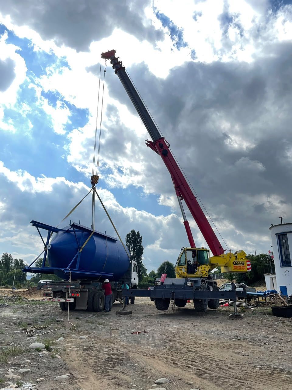 Grúa levantando un remolque azul para embarcaciones en un terreno de grava bajo un cielo nublado
