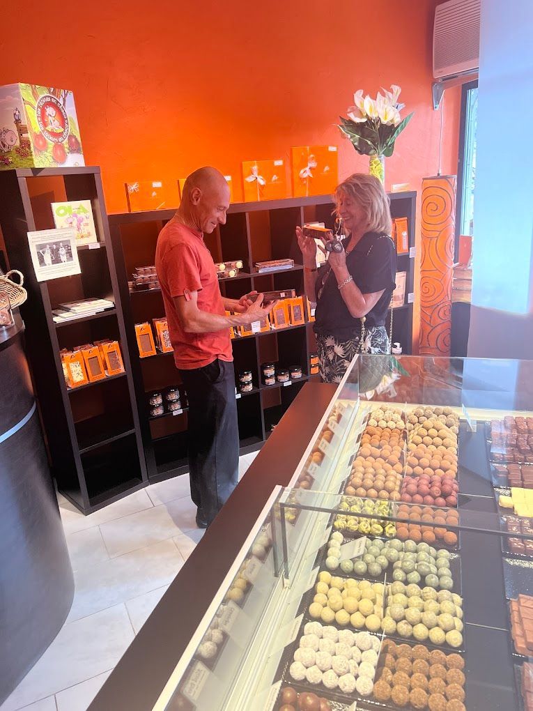 Un homme et une femme font leurs achats dans une chocolaterie. L'homme tient des articles, la femme sourit derrière la vitrine de friandises. Murs orange.
