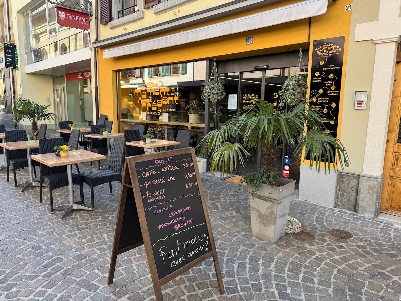 Extérieur d'un restaurant avec terrasse, façade jaune et tableau des menus sur une rue pavée.