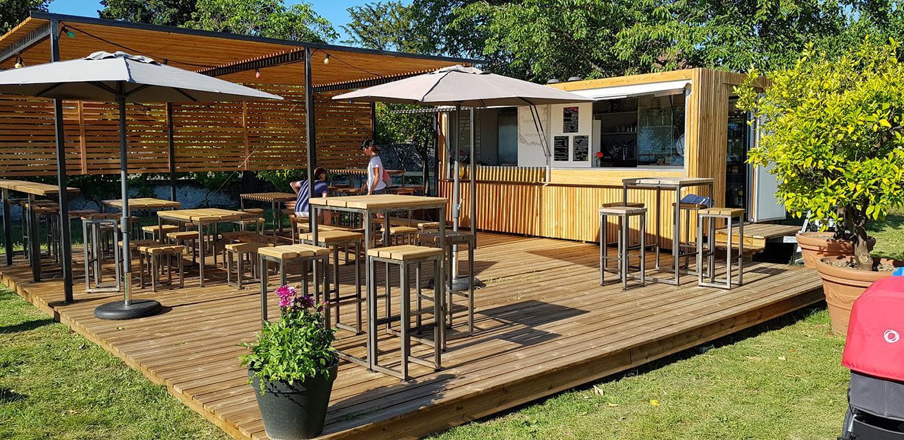Bar extérieur et espace détente avec terrasse en bois, parasols et bâtiment de type conteneur.