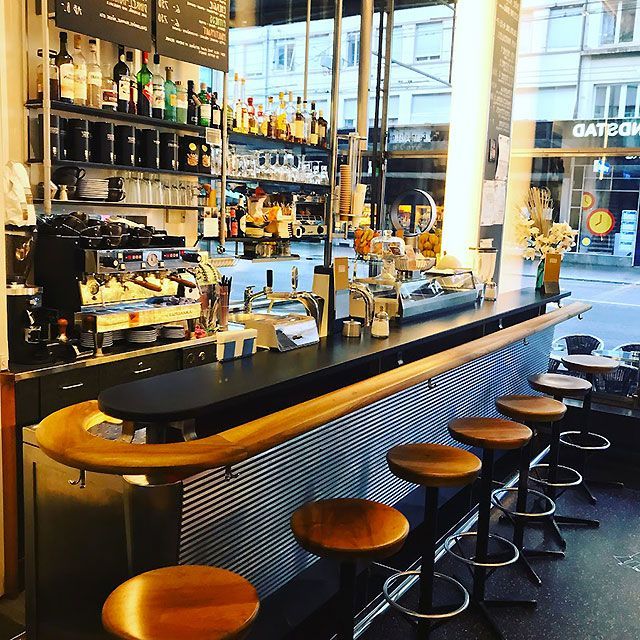 Intérieur d'un café avec des tabourets de bar le long d'un comptoir en bois incurvé, des bouteilles sur des étagères et une machine à café.