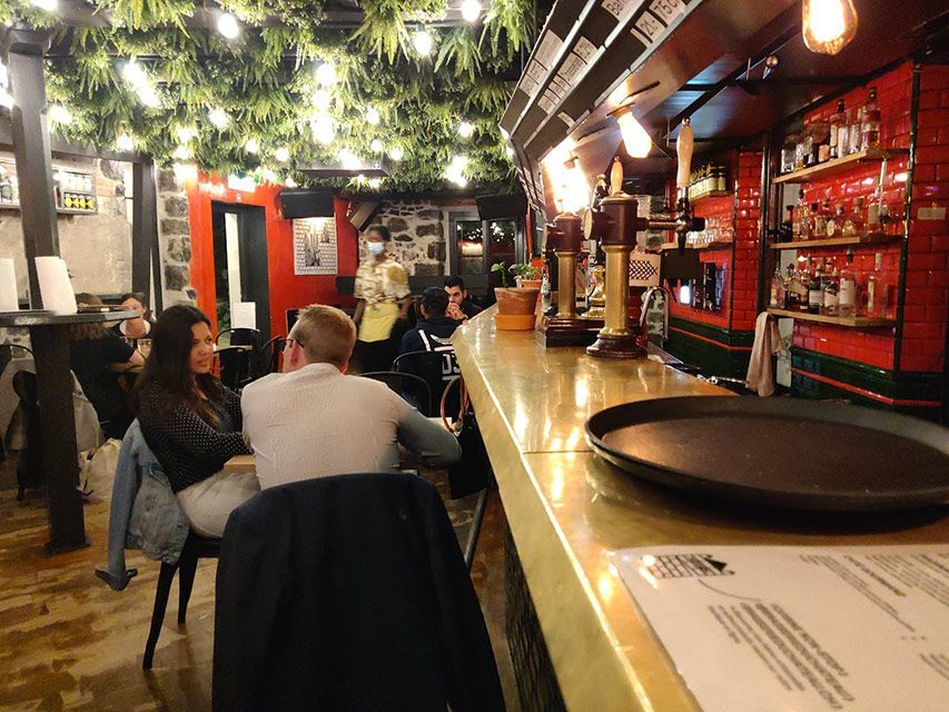 Intérieur d'un bar avec des clients assis au comptoir et aux tables. Des lumières vives éclairent le bar, laissant apparaître les boissons et les verres.