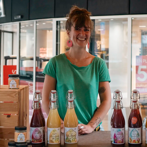Une femme sourit derrière des bouteilles de boissons dans un magasin, vêtue d'une chemise verte.