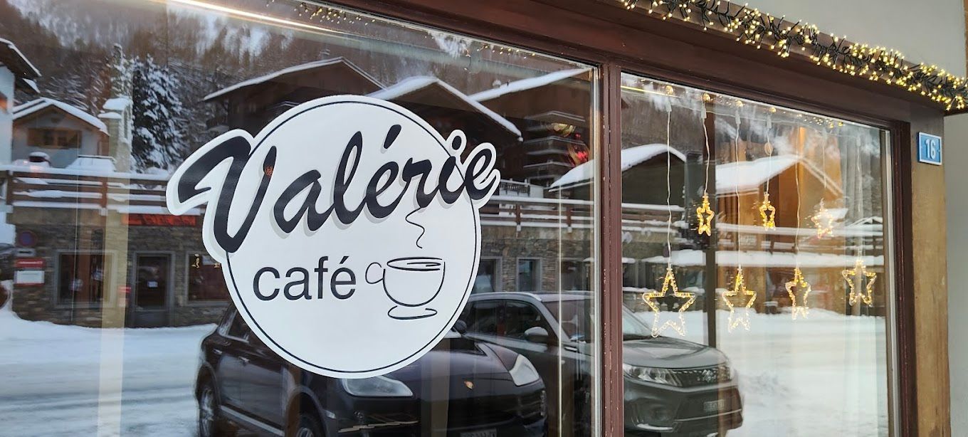 L'enseigne « Valerie Café » est visible dans une vitrine, laissant apparaître le reflet d'un village de montagne enneigé. Des voitures sont visibles devant la vitrine.