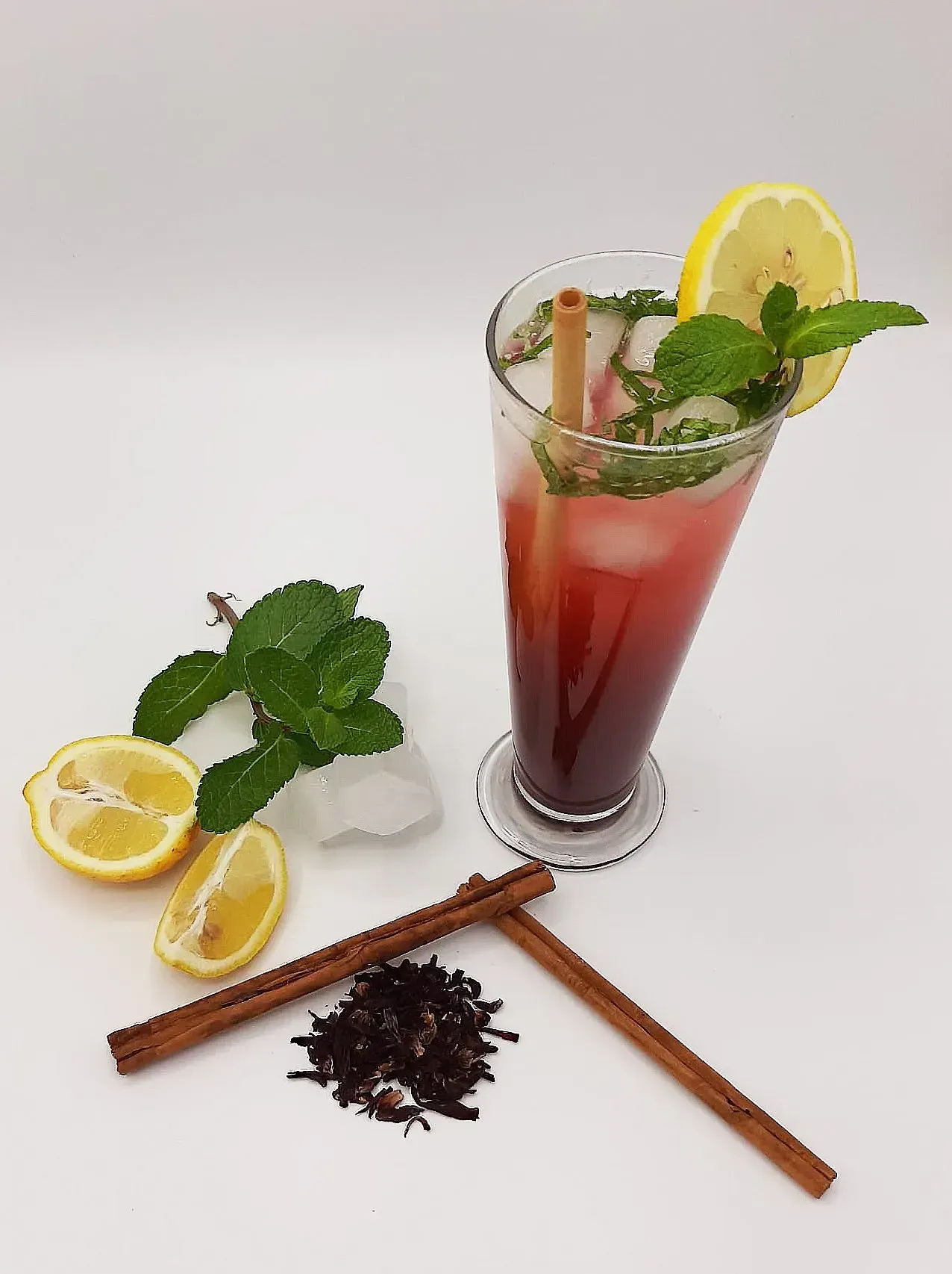 Verre de boisson rouge avec glace, menthe, citron, bâtonnets de cannelle et feuilles de thé en vrac sur du papier blanc.