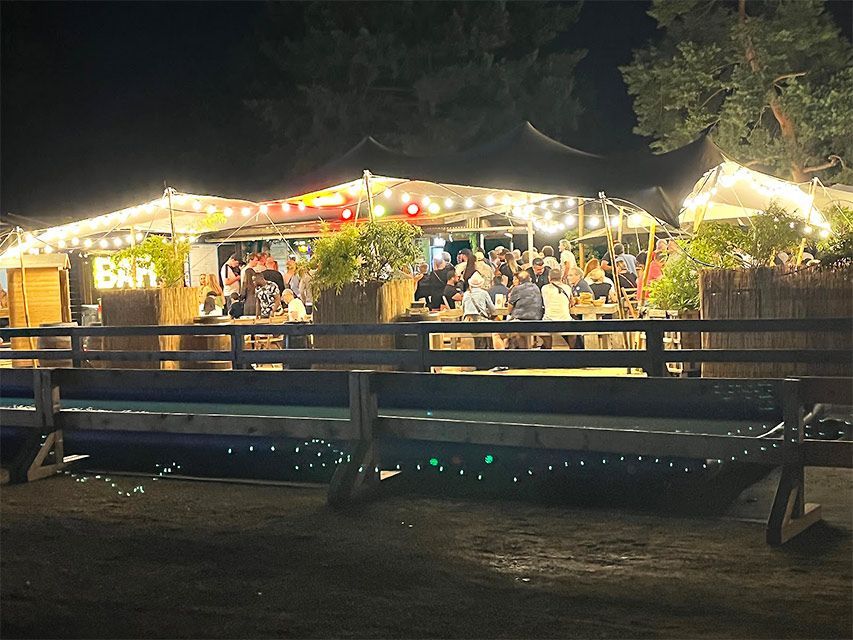 Vue nocturne d'un bar animé en plein air, avec des guirlandes lumineuses, une terrasse en bois et des gens qui discutent.