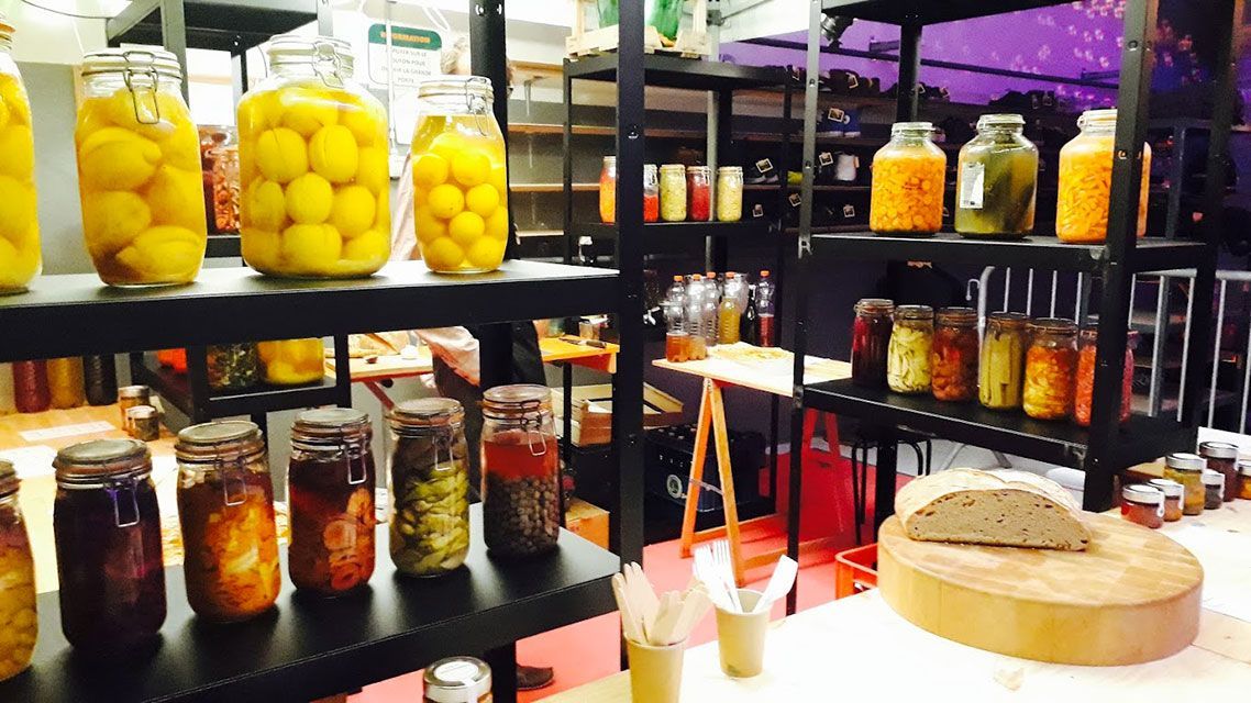 Des étagères remplies de bocaux de conserves, présentées dans une boutique avec du pain et un comptoir en bois.