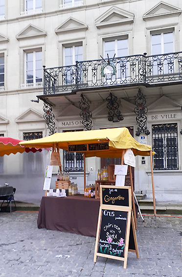 Étal de marché devant un bâtiment classique avec un balcon orné ; auvent, nappe marron, ardoise.