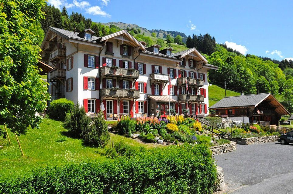 Hôtel dans un paysage montagneux, bâtiment blanc aux volets et balcons rouges, végétation luxuriante.