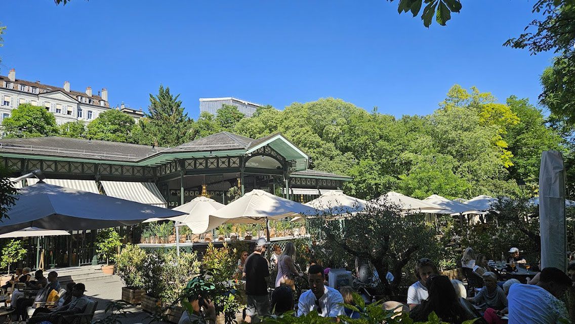 Restaurant en plein air avec parasols, arbres et clients ; bâtiments en arrière-plan sous un ciel bleu.