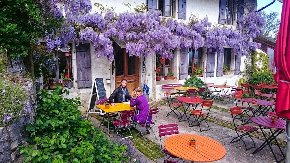 Café en plein air avec glycine violette, tables et clients assis.