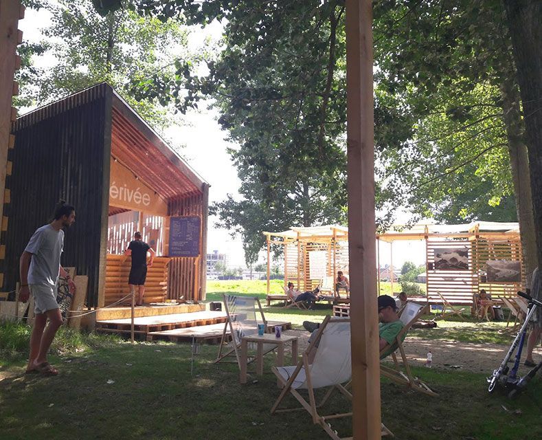 Des personnes se détendent en plein air près de structures en bois. Une personne se tient près d'un kiosque à panneaux. Des arbres sont visibles en arrière-plan.