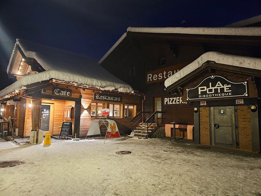 Bâtiment en bois enneigé avec des enseignes « Café », « Restaurant » et « Pizzeria/Bar » la nuit.