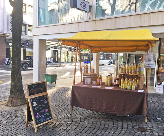 Étal de marché en bord de rue, sous un auvent jaune. Nappe marron, divers flacons et bocaux de produits exposés. Panneau « Ouvert ».
