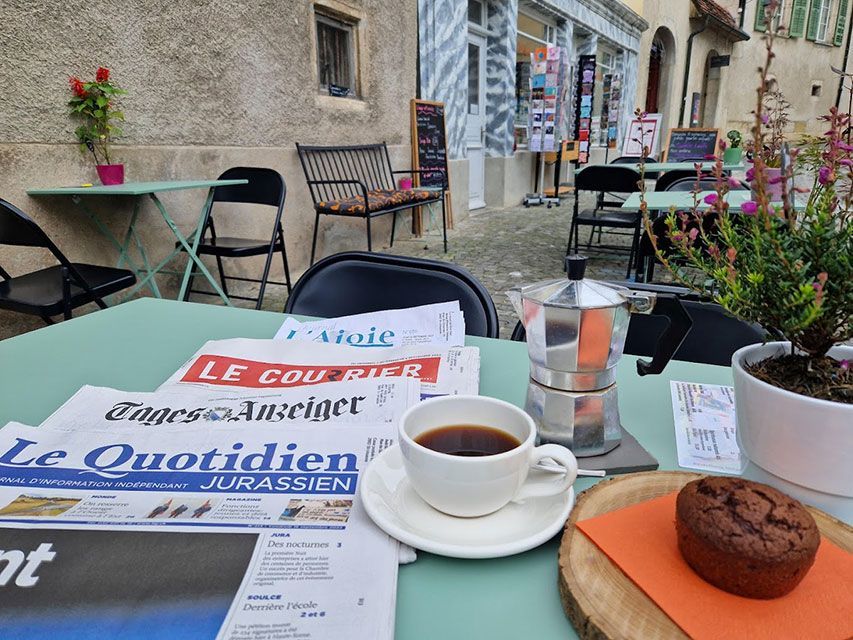 Tasse de café, journal et muffin sur une table de café. Vue extérieure avec des chaises et des bâtiments en arrière-plan.