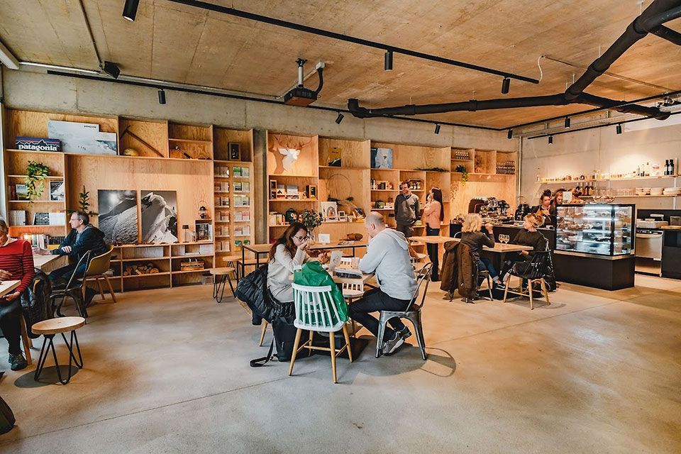 Intérieur d'un café avec des clients attablés. Des étagères en bois longent le mur, présentant divers objets. Sol et plafond en béton.
