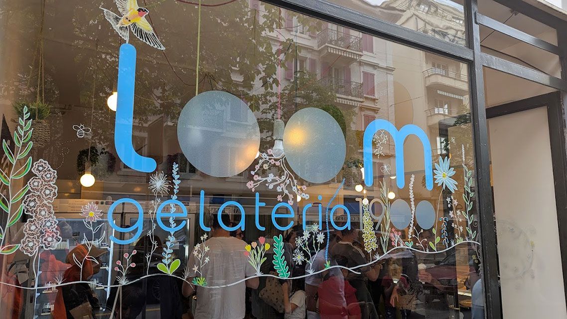 « Façade de Loom Gelato avec de grandes lettres bleues sur verre. On aperçoit des personnes à l'intérieur et les reflets des bâtiments. »