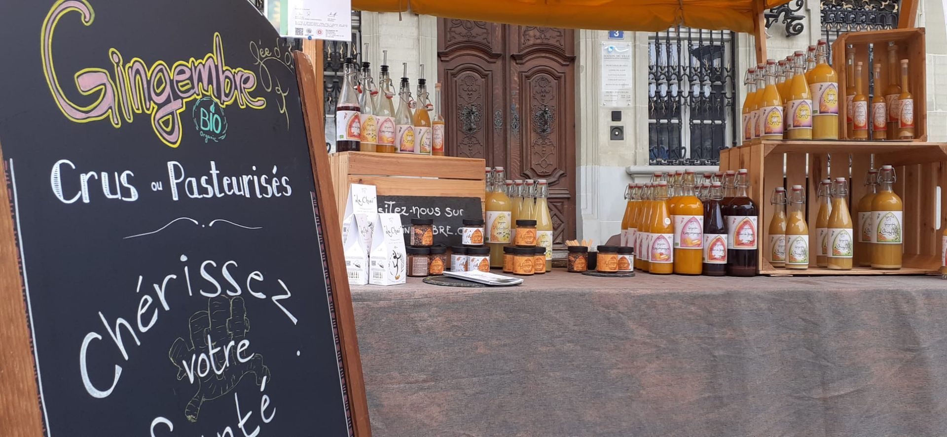 Un étal de marché où sont exposés divers bocaux et bouteilles de produits. On aperçoit une ardoise.