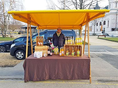 Un homme vend du jus de fruits sur un étal de marché en plein air, sous un auvent jaune. Bouteilles et gobelets sont posés sur une table recouverte d'une nappe marron.
