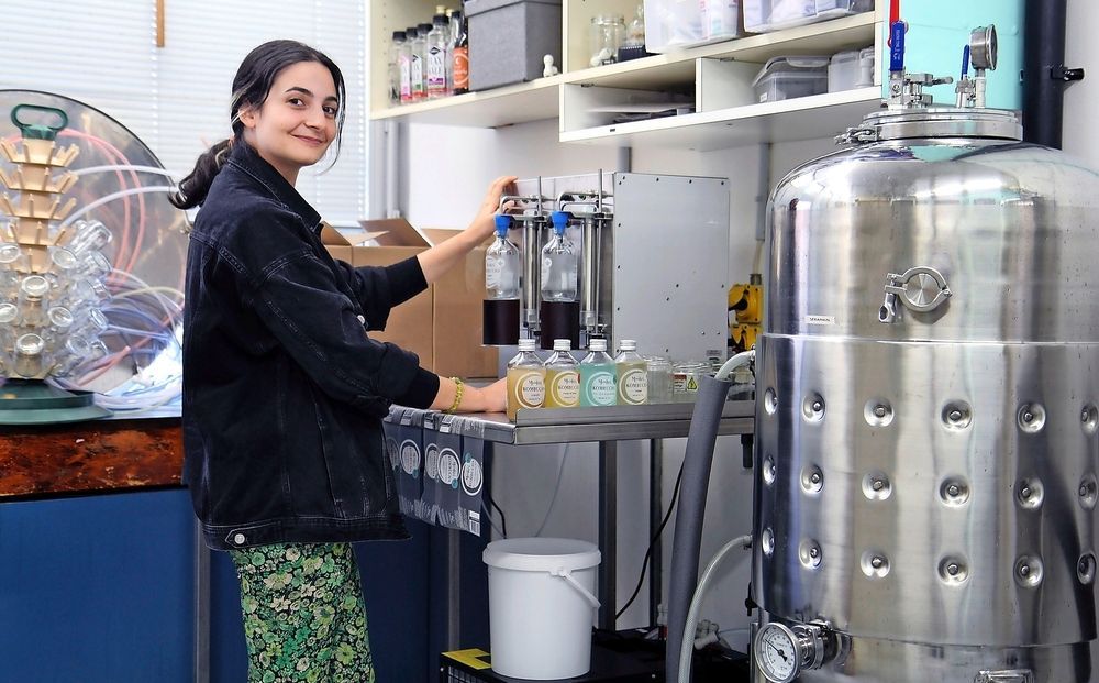 Une femme en blouse blanche remplit des flacons de liquide en souriant. Des cuves et des équipements en acier inoxydable sont visibles en arrière-plan.