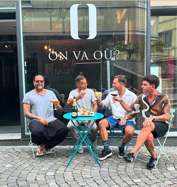 Quatre personnes trinquent devant un bar orné du logo « O ». Elles sont assises à une table turquoise dans une rue pavée.