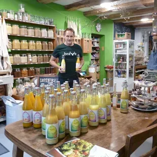 Un homme, tenant des boissons dans un magasin, est entouré de bouteilles de jus jaune posées sur une table en bois. Des rayons de produits alimentaires sont visibles en arrière-plan.